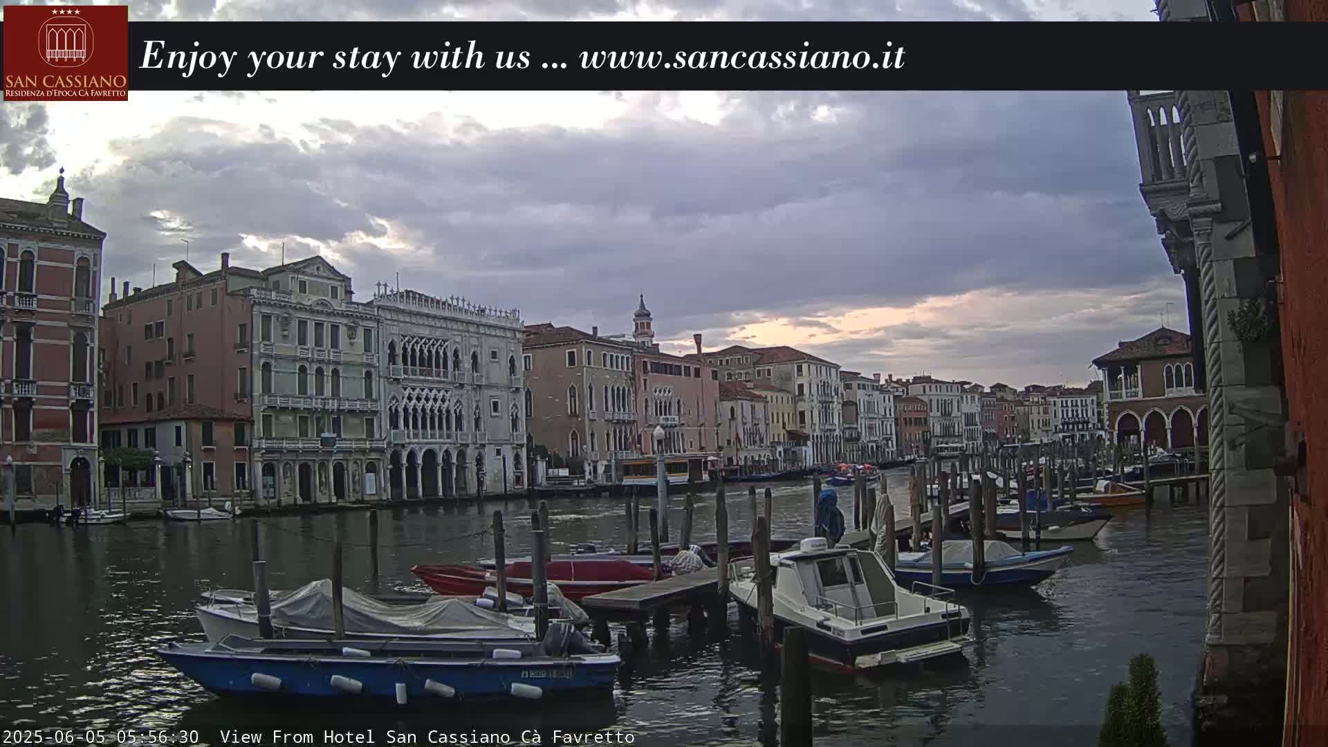 A cloudy sunrise over a Venetian canal, showing several boats moored along the waterfront, with buildings lining the canal.