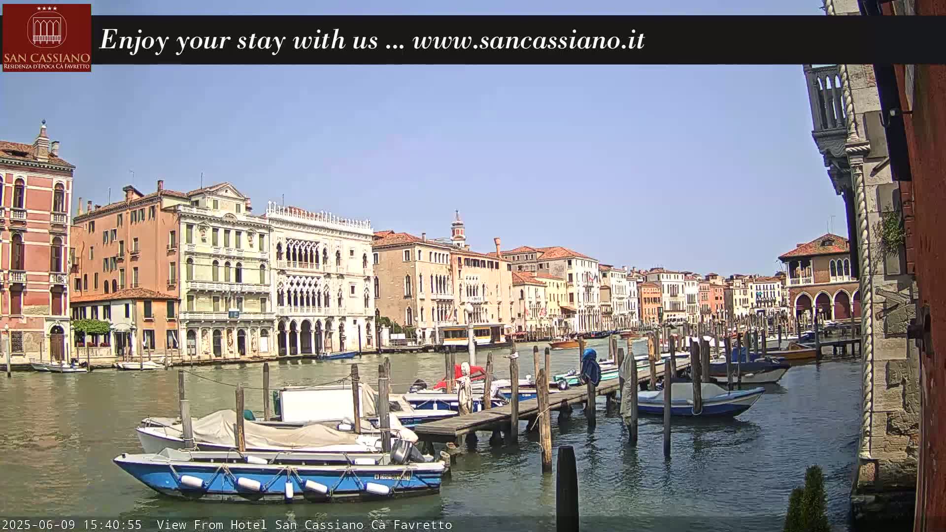 A sunny day shows a canal in Venice, Italy, lined with historic buildings and several boats moored at a dock.