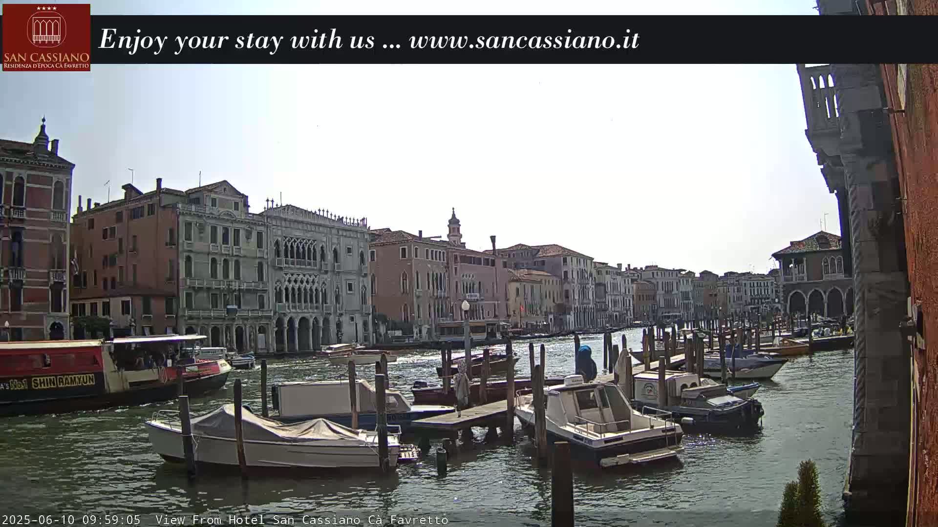 A sunny day in Venice shows a canal lined with historic buildings, numerous boats docked along the waterway, and a larger boat in the foreground.