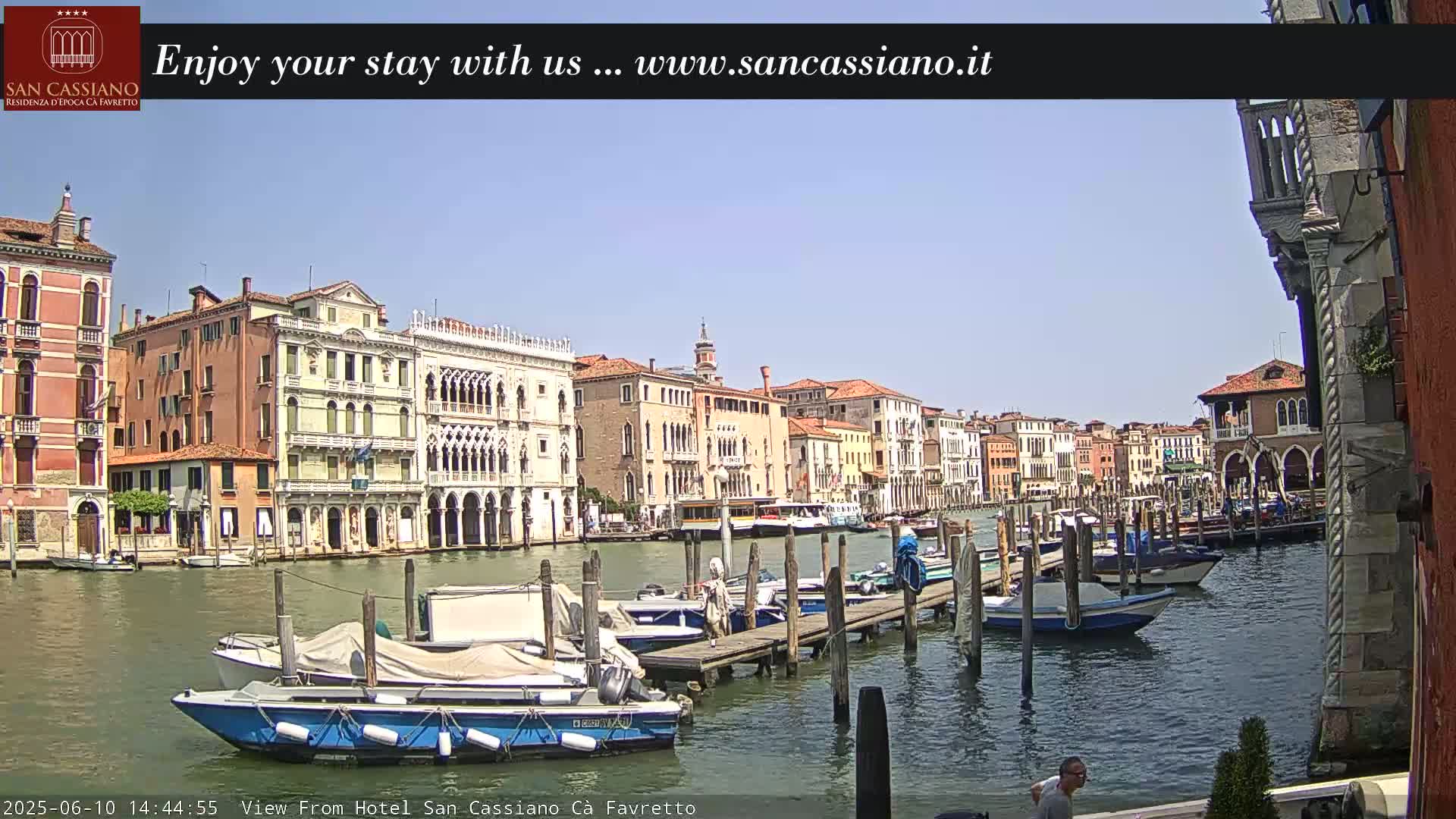 A sunny day in Venice shows a canal with several boats moored at a dock, alongside buildings lining the waterway.