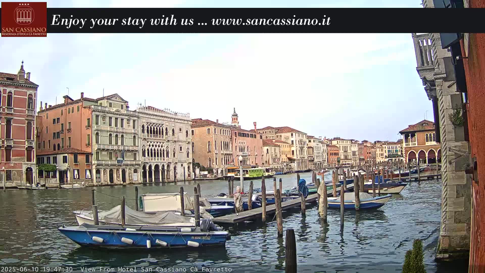A canal in Venice, Italy, with numerous boats moored along a wooden dock, under an overcast sky.