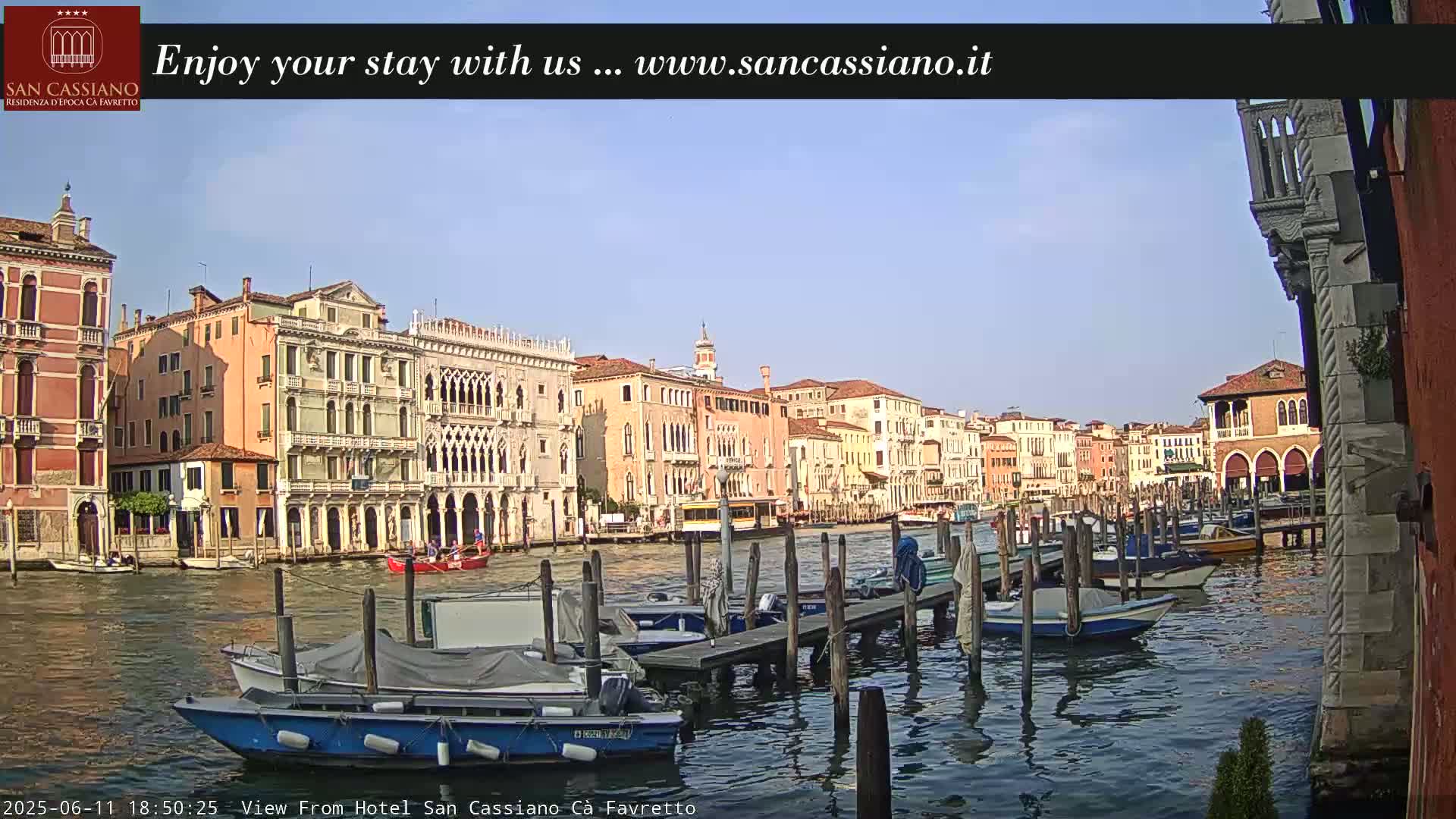 A sunny Venetian canal scene shows colorful buildings lining the waterway, with several boats moored at a dock.