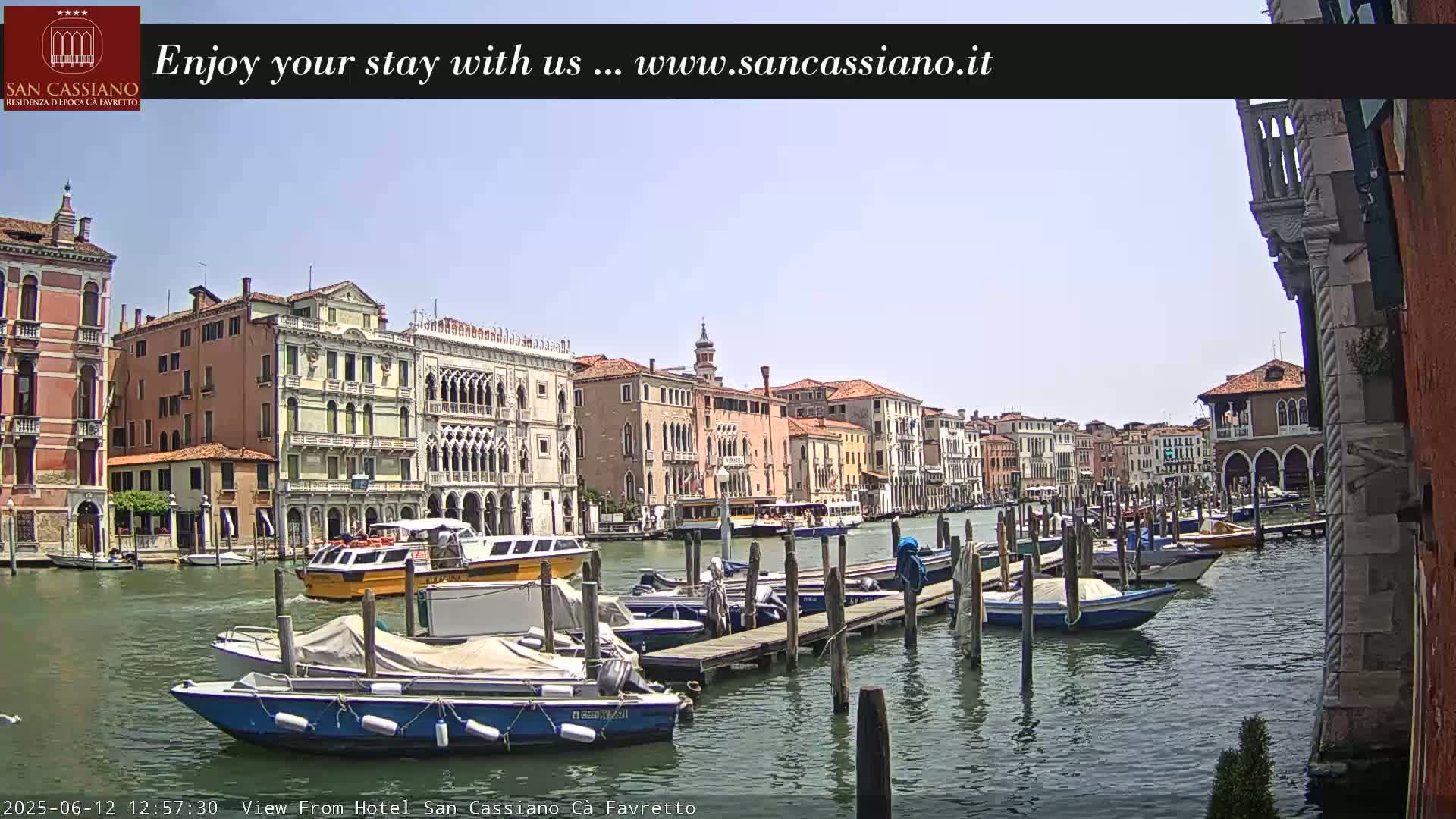 A sunny day in Venice shows a canal lined with colorful buildings, boats moored at docks, and a few watercraft on the waterway.