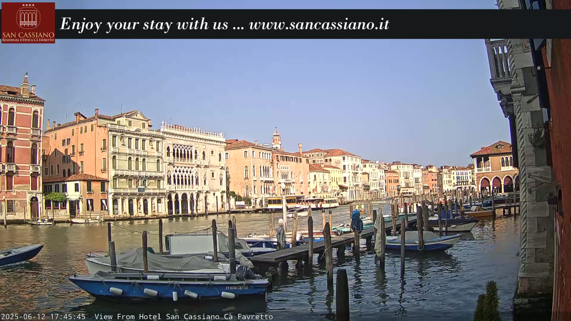 A sunny Venetian canal scene shows several moored boats along a dock in front of colorful buildings.
