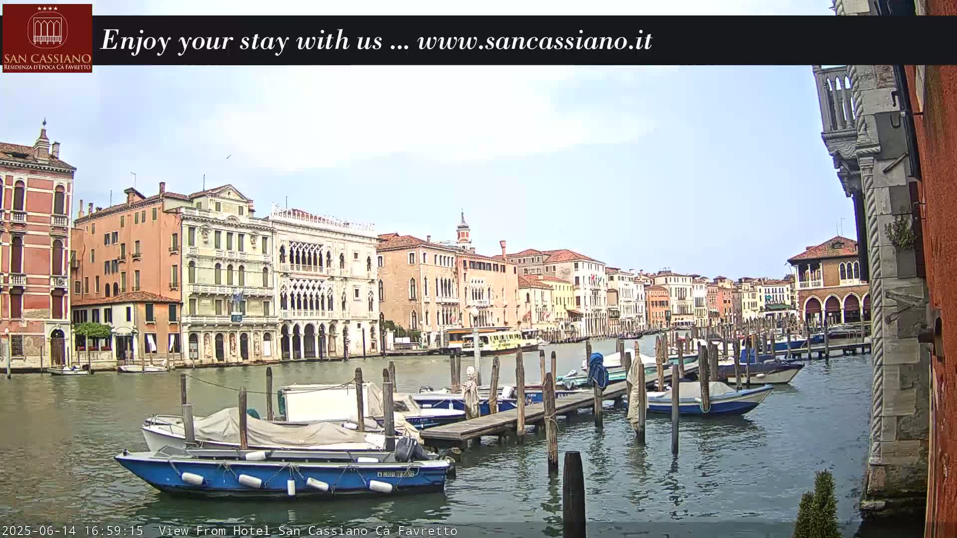 A canal in Venice, under a partly cloudy sky, is lined with colorful buildings and numerous boats moored at wooden docks.