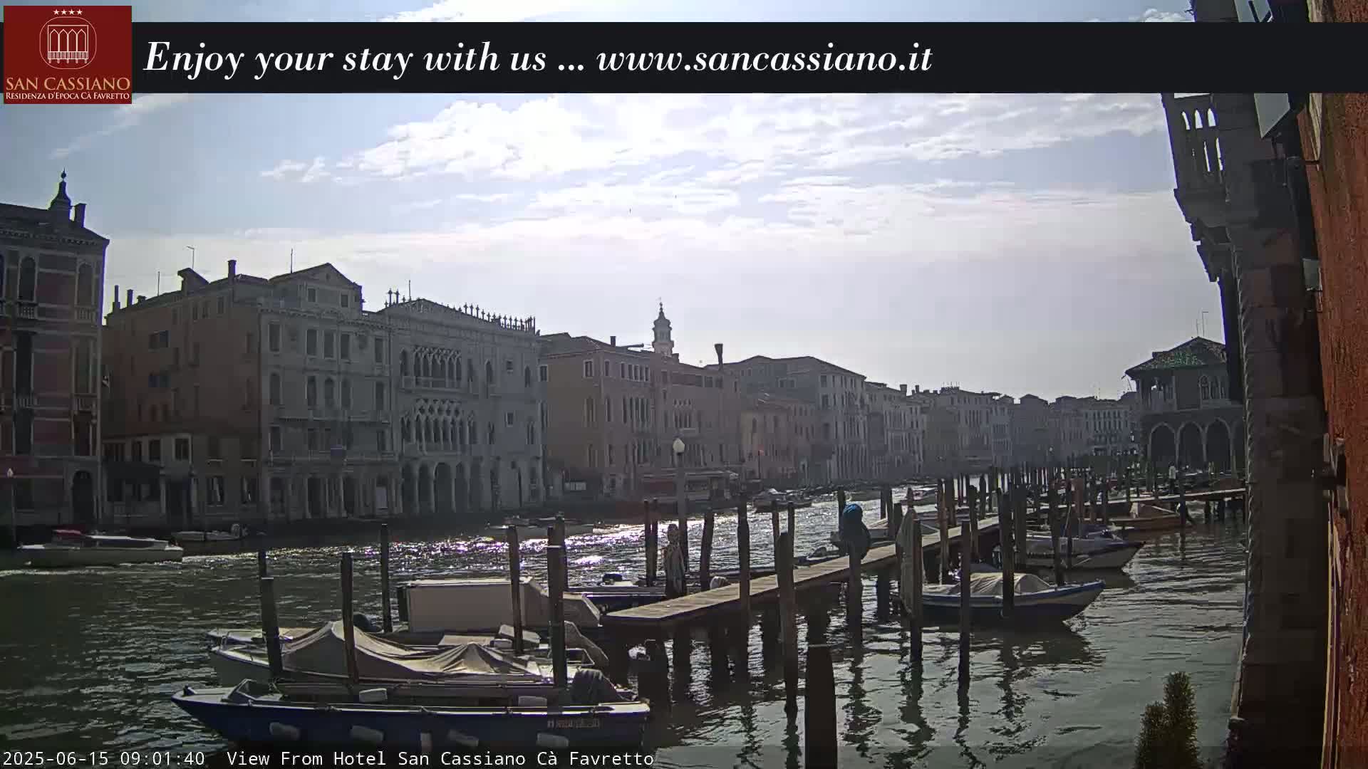 A sunny day in Venice shows a canal lined with historic buildings, boats docked at a wooden pier, and a few people standing on the pier.