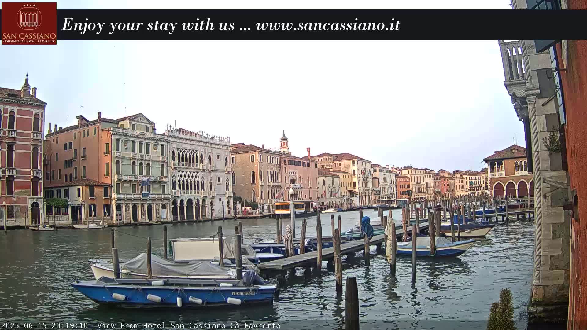 A canal in Venice, Italy, shows many boats moored at wooden docks in front of several multi-story buildings under a mostly cloudy sky.