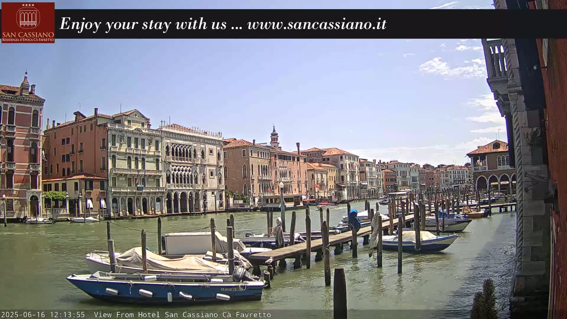 A sunny day in Venice shows several boats moored at a dock along a canal, with many buildings lining the waterway.