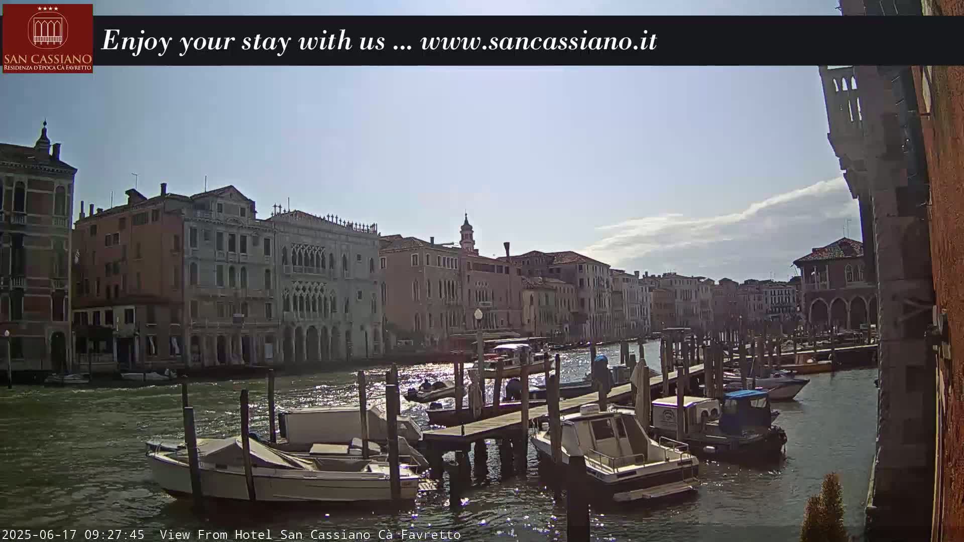 A sunny day in Venice shows several boats moored at a dock along a canal lined with historic buildings.