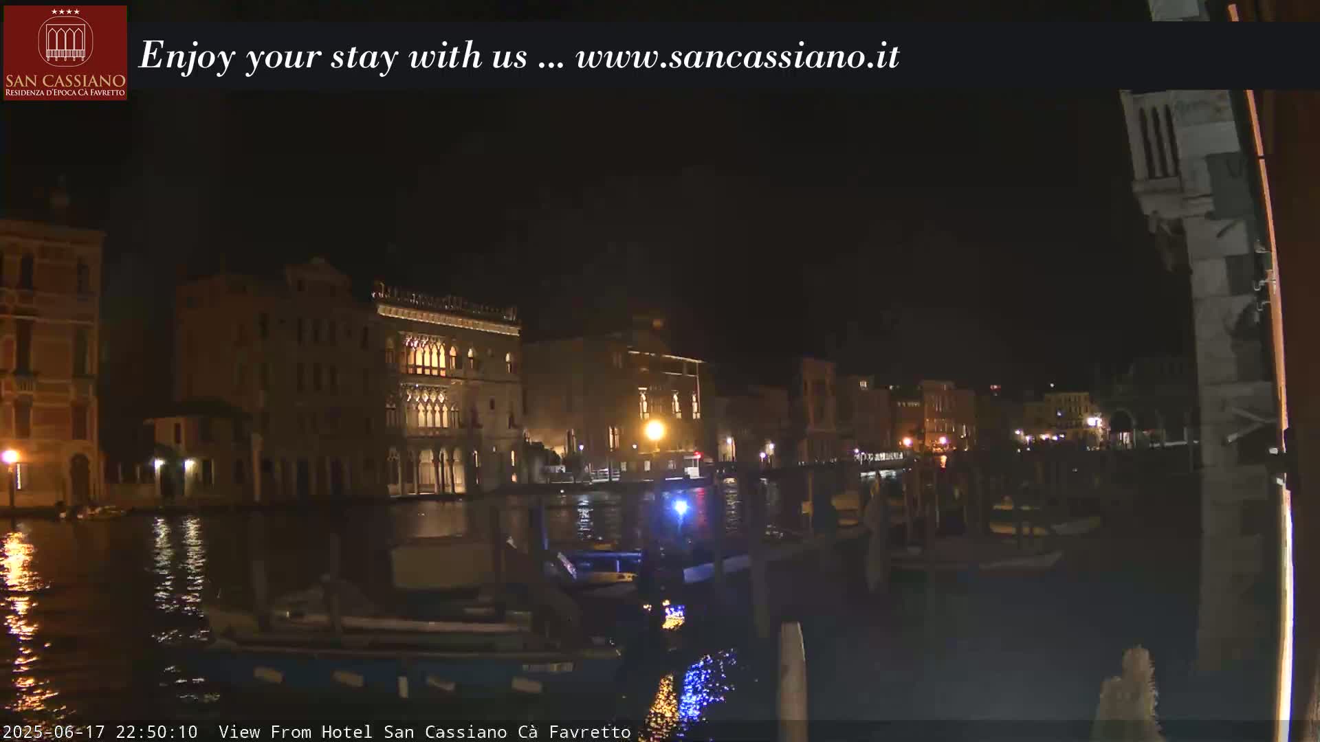 A nighttime view of a canal in Venice, with illuminated buildings lining the waterway and boats moored along the edge.