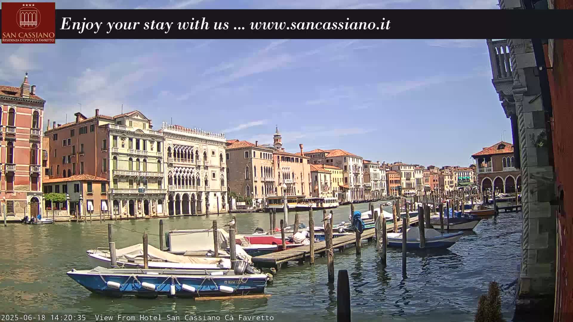 A sunny day in Venice shows colorful buildings lining a canal, with several boats moored at a dock.