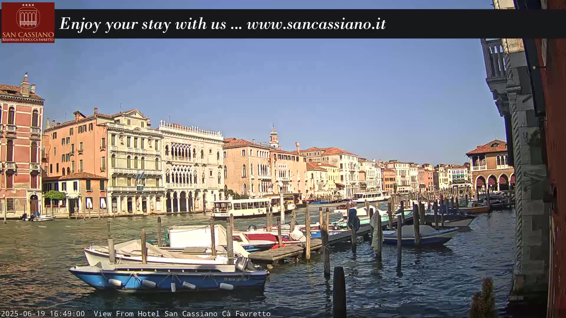 Under a clear blue sky, a Venetian canal is lined with colorful buildings, boats docked along a pier, and a water bus traveling along the waterway.