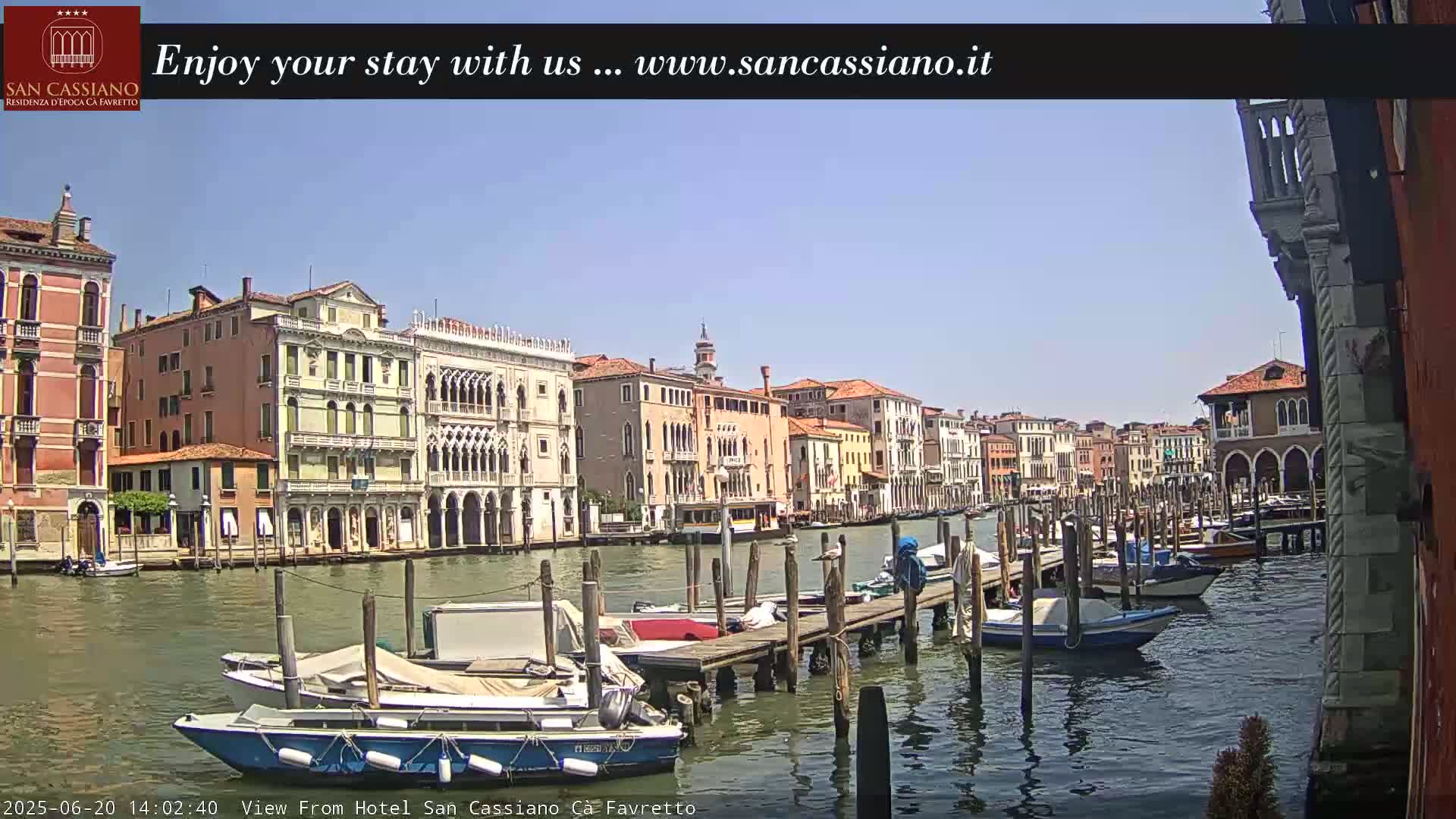 A sunny day in Venice shows a canal lined with historic buildings and numerous boats moored at wooden docks.
