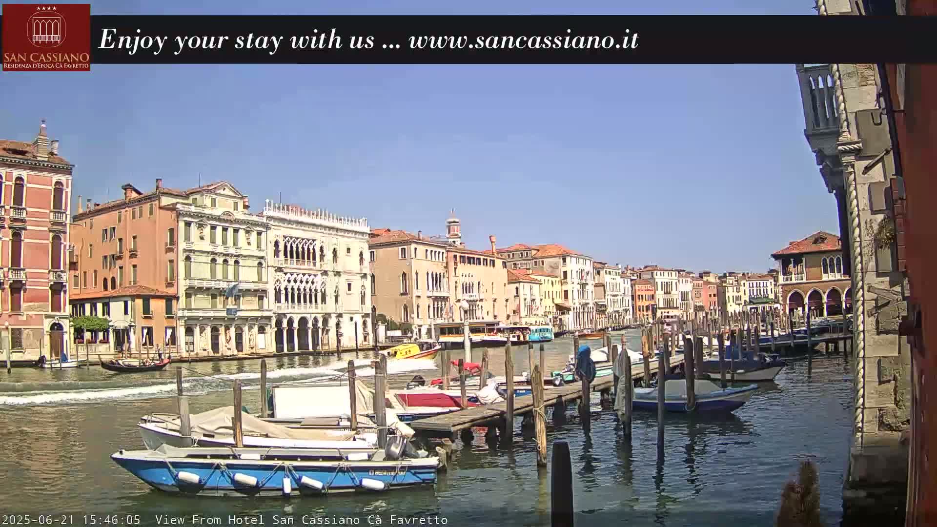 A sunny day in Venice shows several boats moored at a dock along a canal, with colorful buildings lining the waterway.