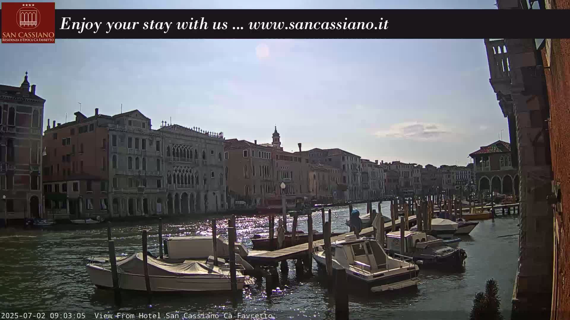 A sunny day in Venice shows a canal lined with buildings, boats docked at piers, and several motorboats on the water.