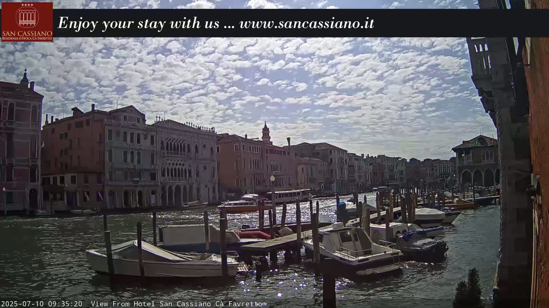 A canal in Venice, under a partly cloudy sky, is lined with buildings and numerous moored boats.