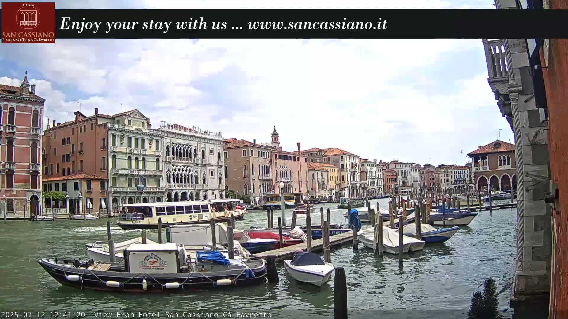 A canal in Venice, under a partly cloudy sky, is lined with buildings and numerous moored boats.