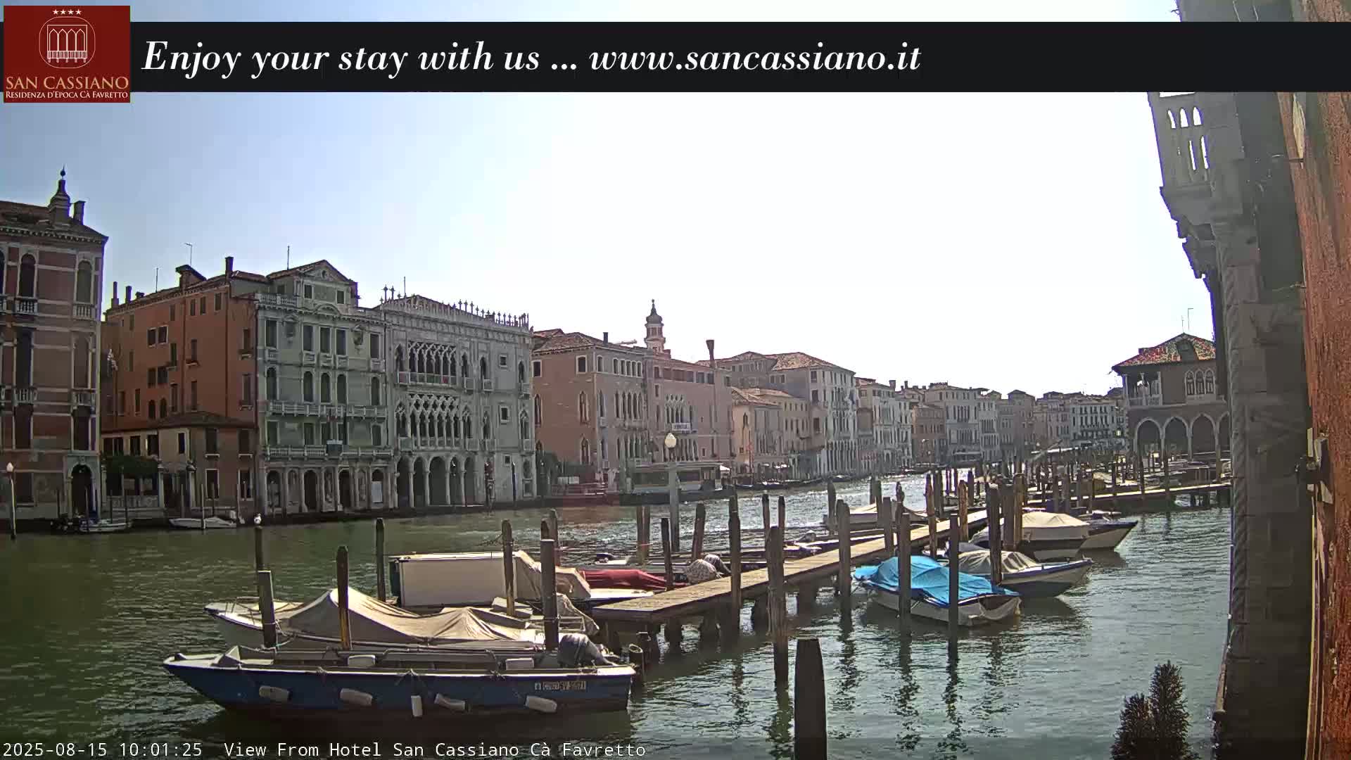A sunny day in Venice shows a canal lined with buildings, boats moored at a dock, and a clear blue sky.