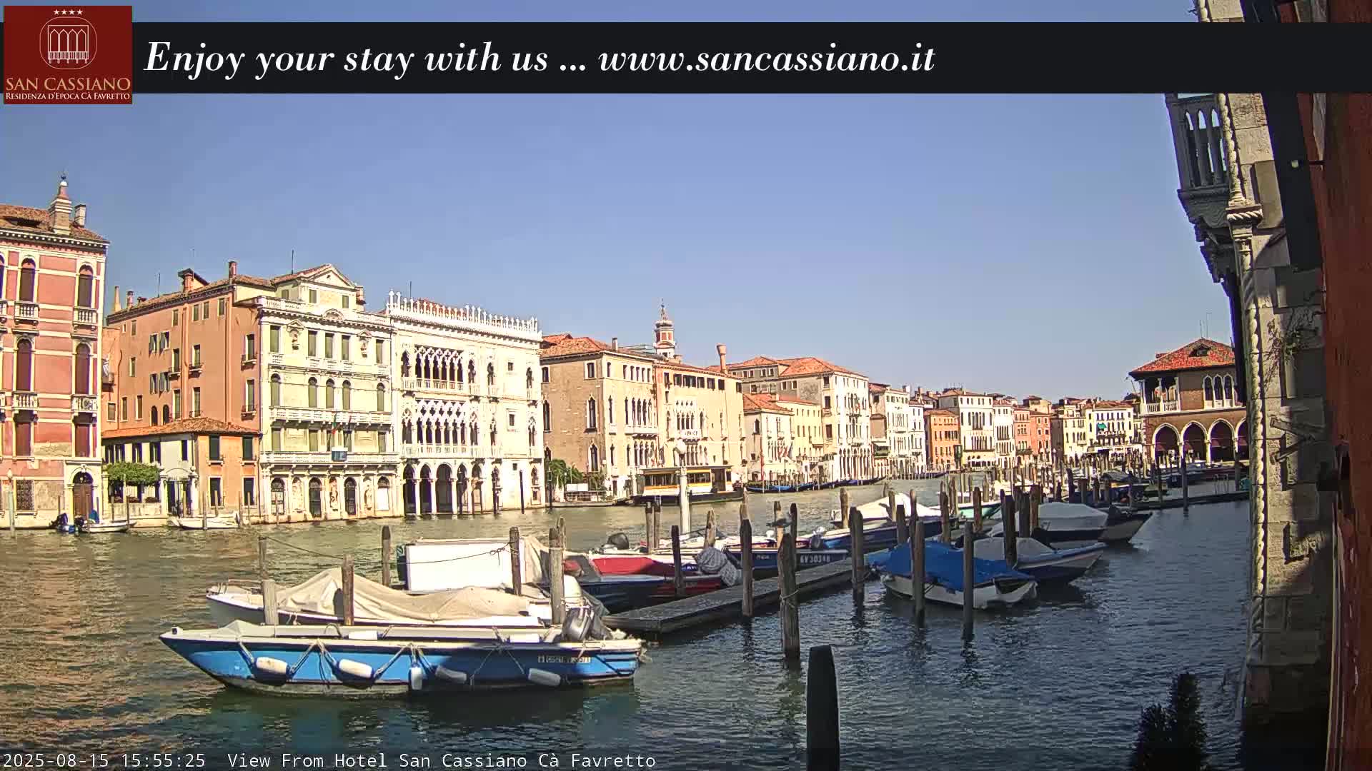 A sunny day in Venice shows a canal lined with buildings, boats moored at a dock, and a clear blue sky.