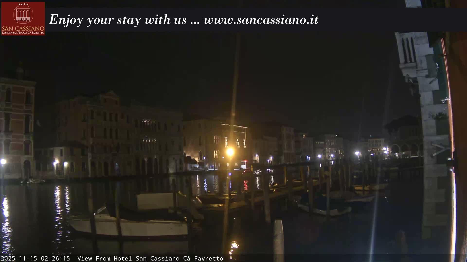 A clear night view of a Venetian canal, featuring historic buildings illuminated by lights, boats moored along the banks, and their reflections shimmering on the water.