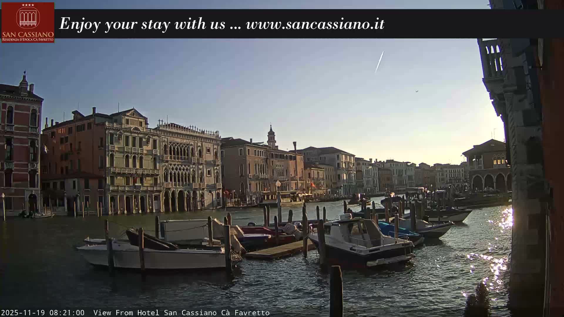 A clear night view of a Venetian canal, featuring historic buildings illuminated by lights, boats moored along the banks, and their reflections shimmering on the water.