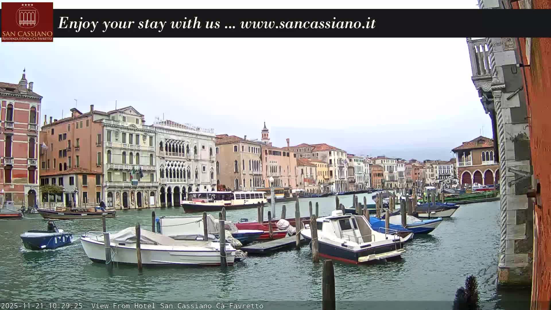 A wide canal flows between densely packed, ornate historic buildings under an overcast sky, with numerous boats, some docked and some moving, dotting the water.