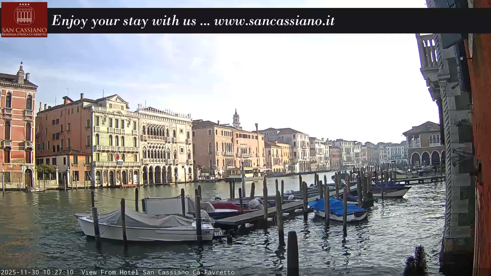 A scenic view of a Venetian canal, lined with ornate historic buildings, features numerous boats docked along wooden posts under a clear sky.