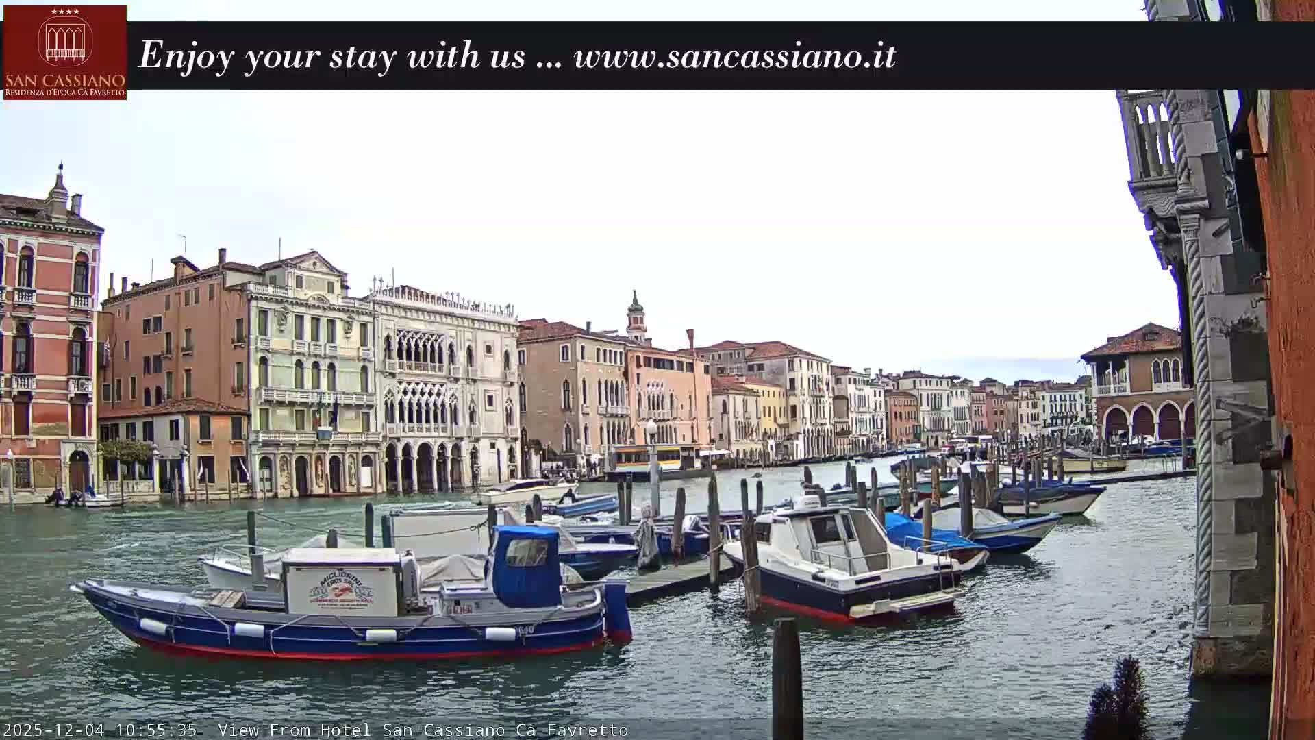 A wide canal in Venice is lined with ornate historic buildings and filled with numerous docked boats under a bright, overcast sky.