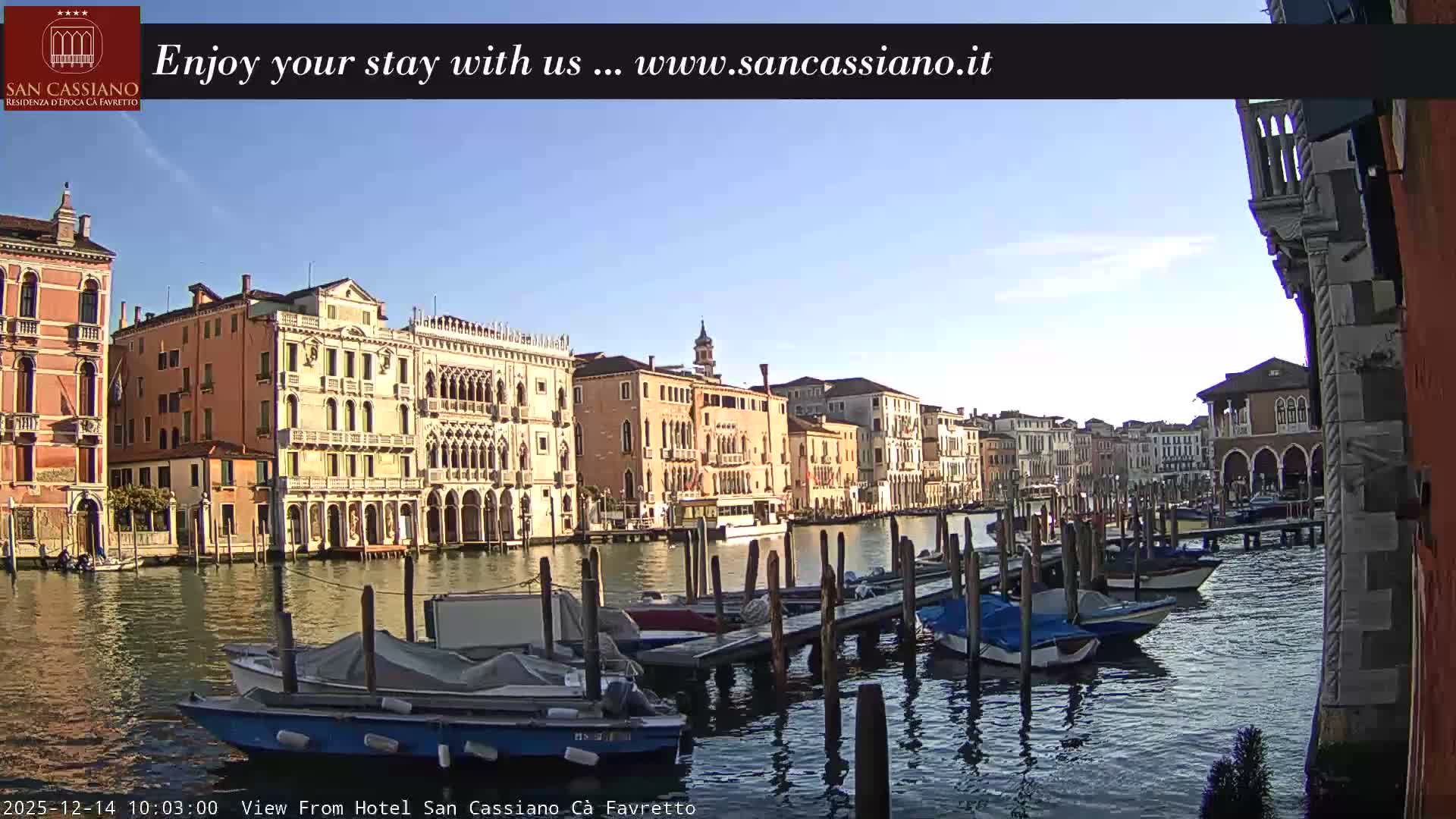 A wide canal in Venice is lined with ornate historic buildings and filled with numerous docked boats under a bright, overcast sky.