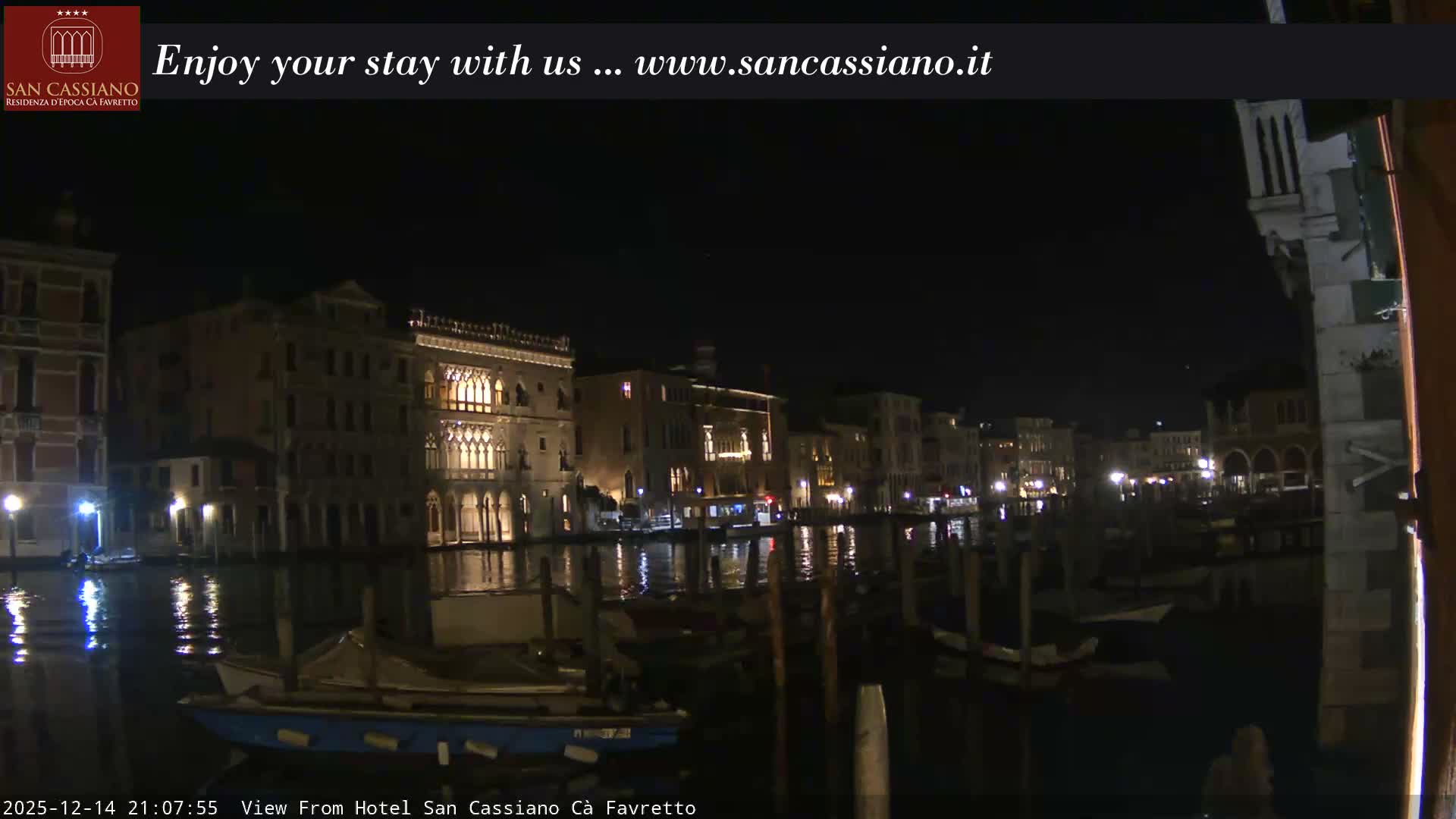 A wide canal in Venice is lined with ornate historic buildings and filled with numerous docked boats under a bright, overcast sky.