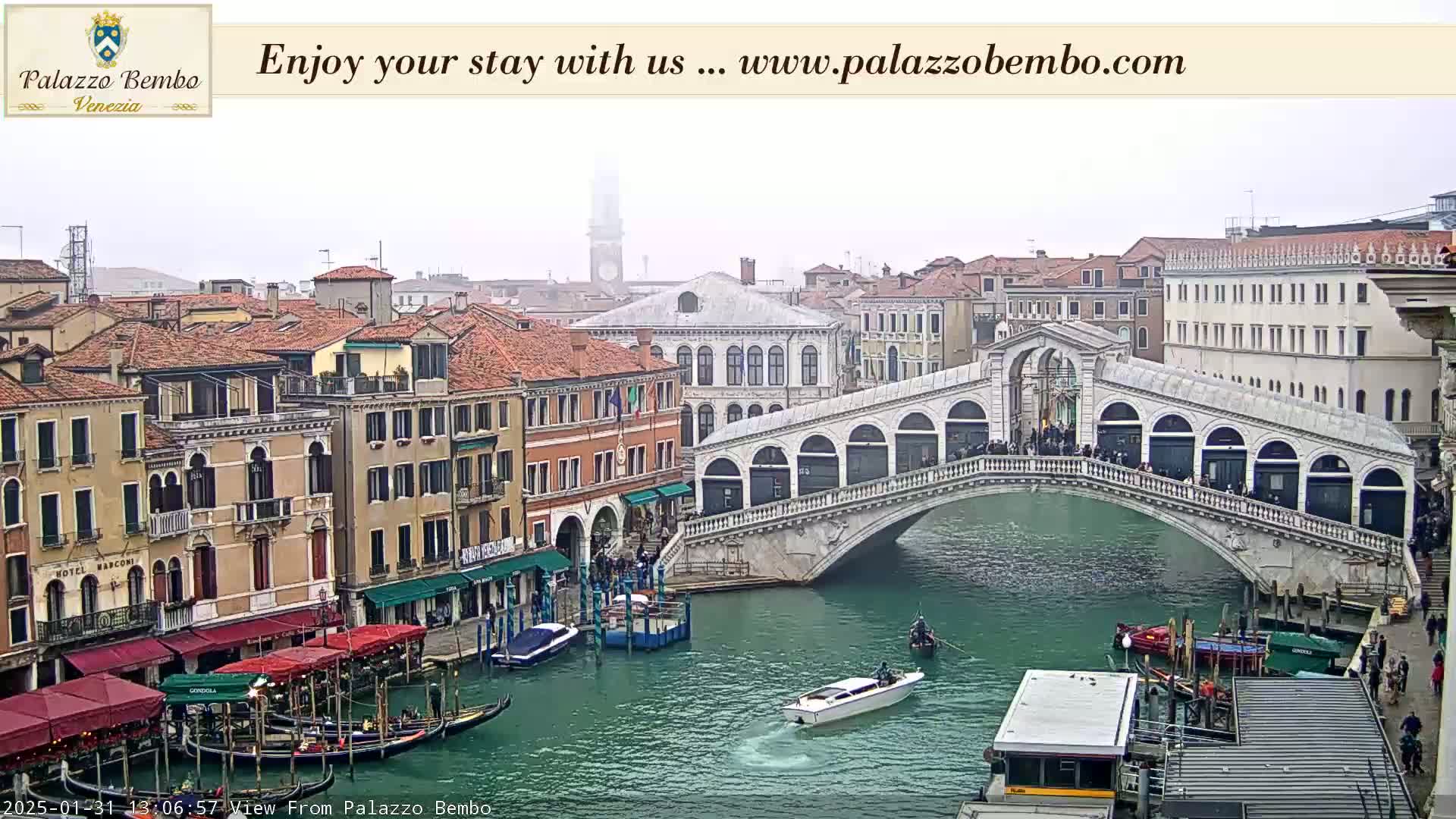 A foggy view of a canal in Venice, showing gondolas, a bridge with people on it, and various buildings along the waterway.
