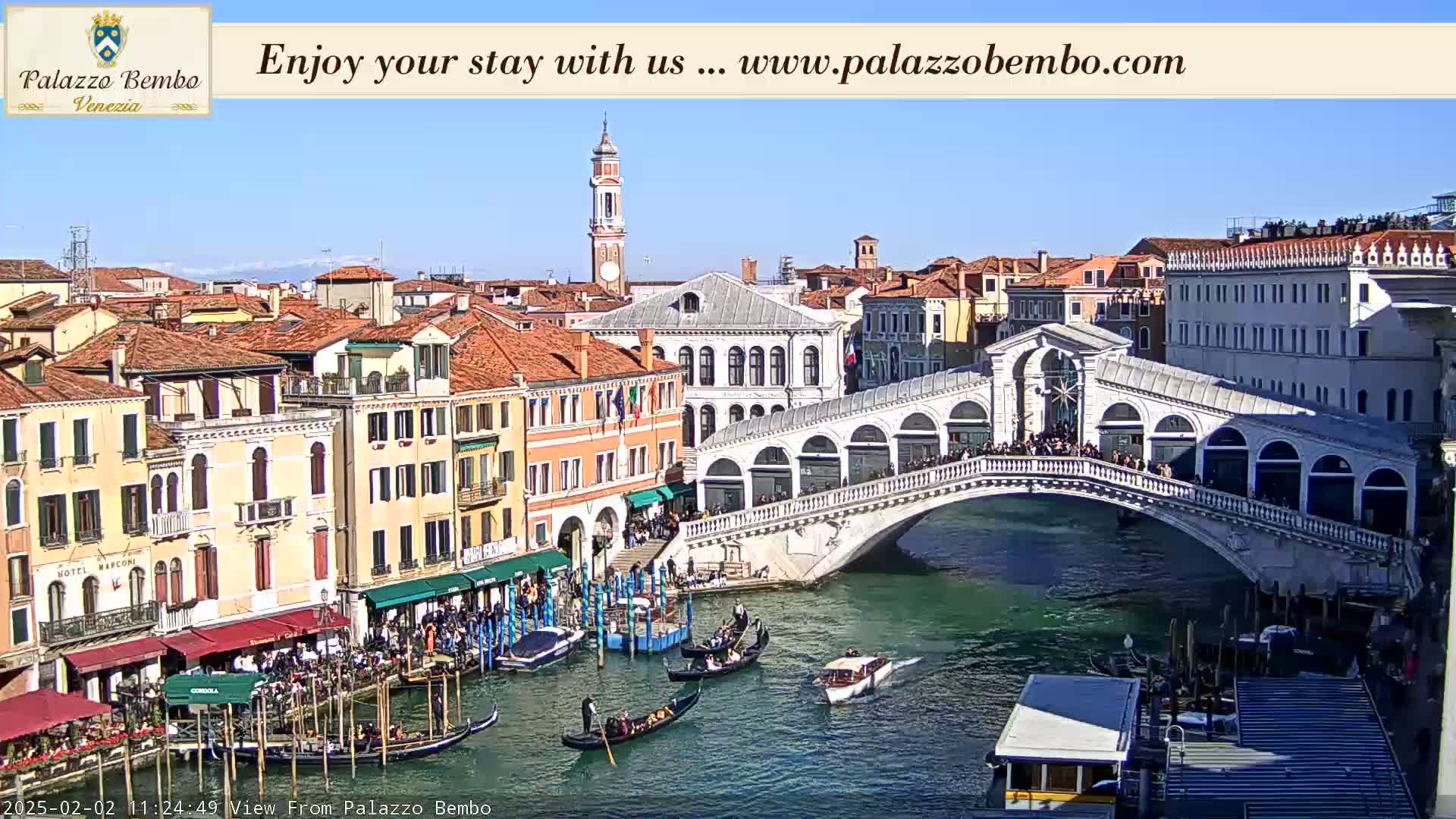A sunny view of a Venetian canal with gondolas, a bridge crowded with people, and numerous buildings lining the waterway.