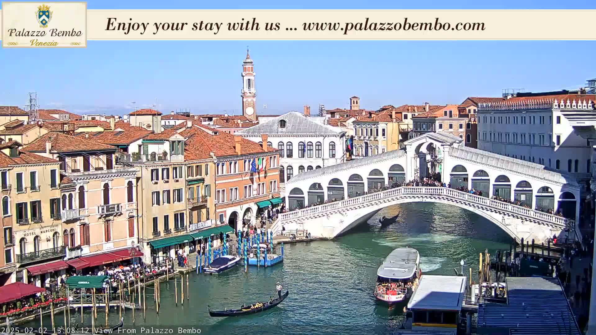 A sunny day in Venice shows a canal with gondolas and boats, a bridge crowded with people, and numerous buildings lining the waterway.