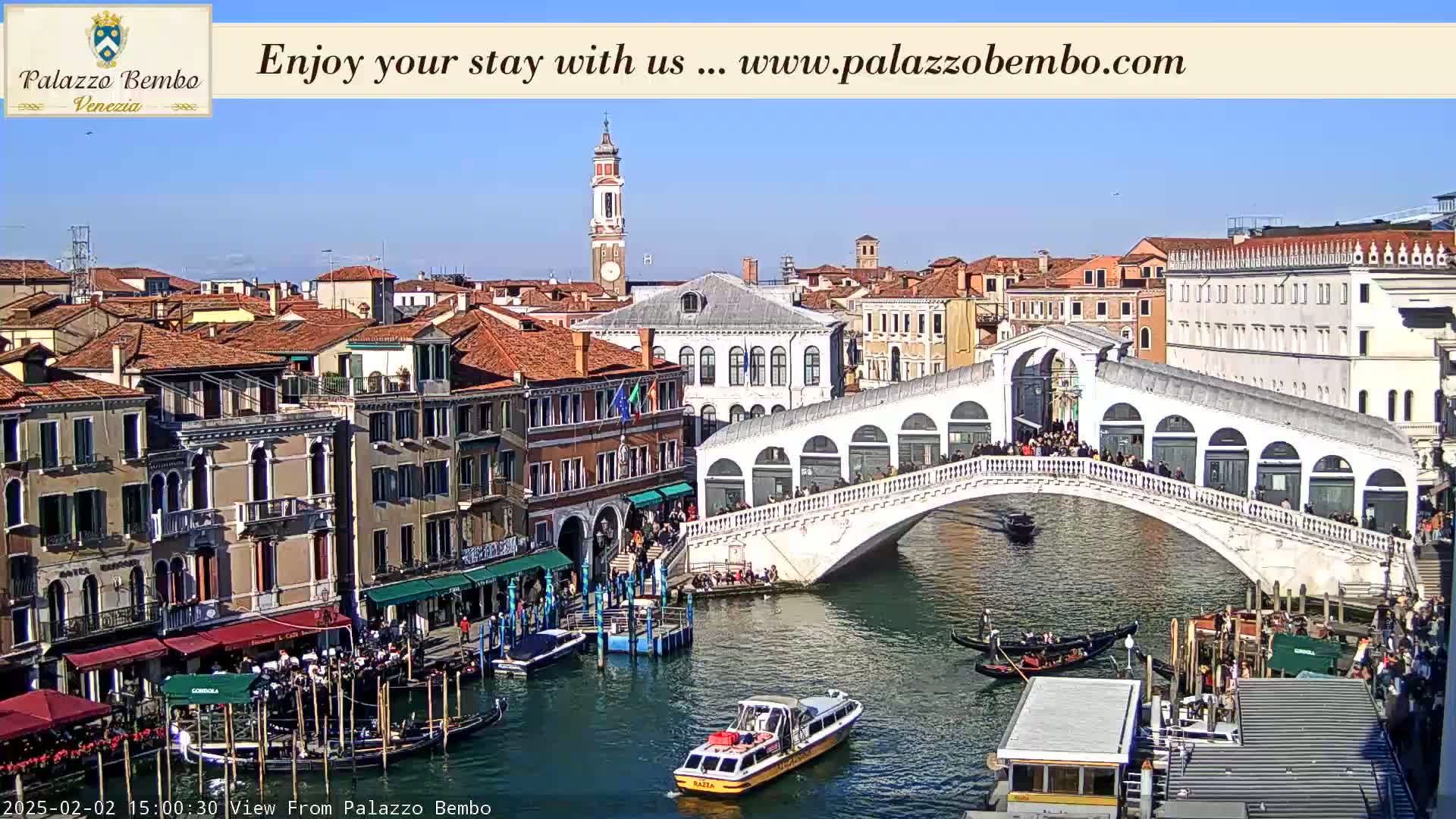 A sunny day in Venice shows a canal with gondolas, a bridge crowded with people, and several buildings lining the waterway.