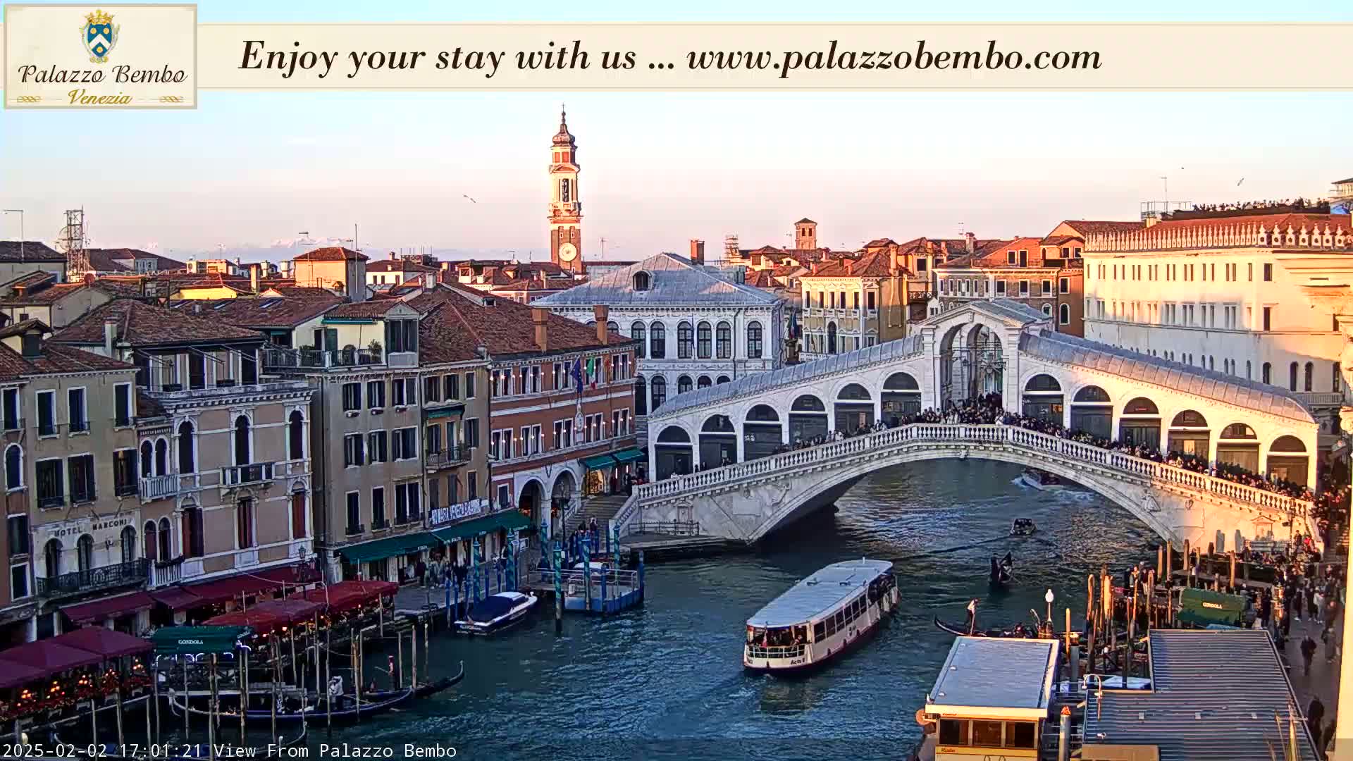 A Venetian bridge crowded with people is seen at sunset over a canal with boats, buildings, and a tall clock tower in the background.