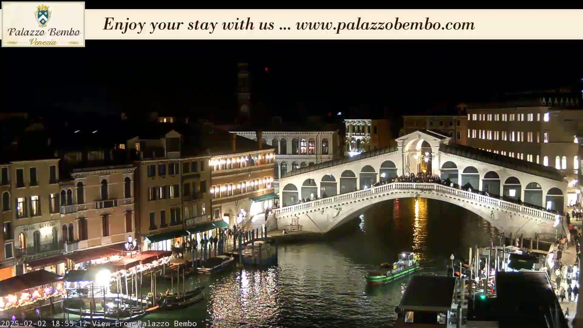 A nighttime view of a canal in Venice, showing a lit bridge with people on it and buildings along the waterway.