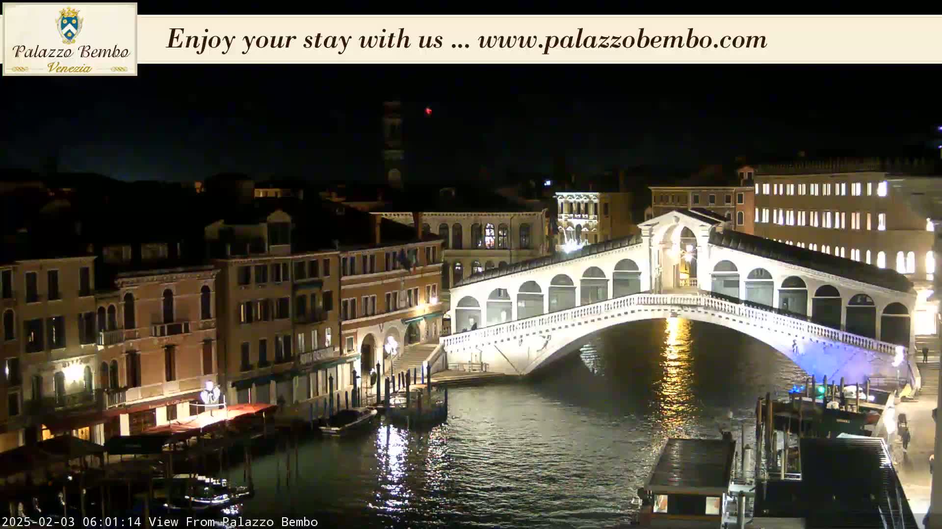 A nighttime view of a canal in Venice, showing a bridge with lights reflecting on the water.