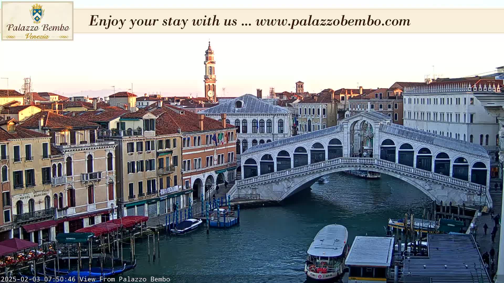 A Venetian canal scene at sunrise shows a stone bridge arching over the water, with colorful buildings lining the waterway, boats moored along the sides, and a few water taxis navigating the canal.