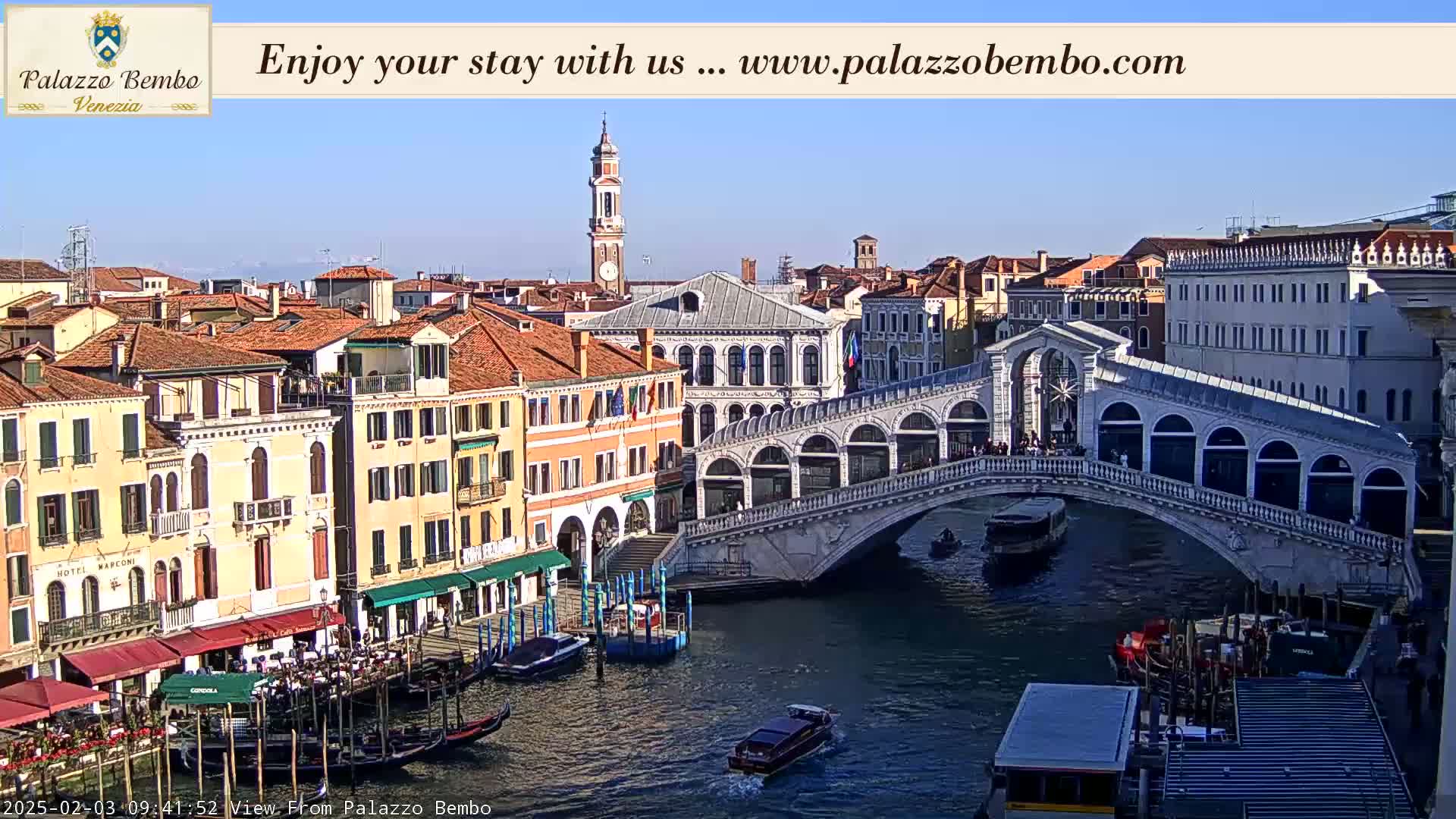 A sunny view of the Rialto Bridge in Venice, with numerous gondolas and boats on the canal and buildings lining the waterway.