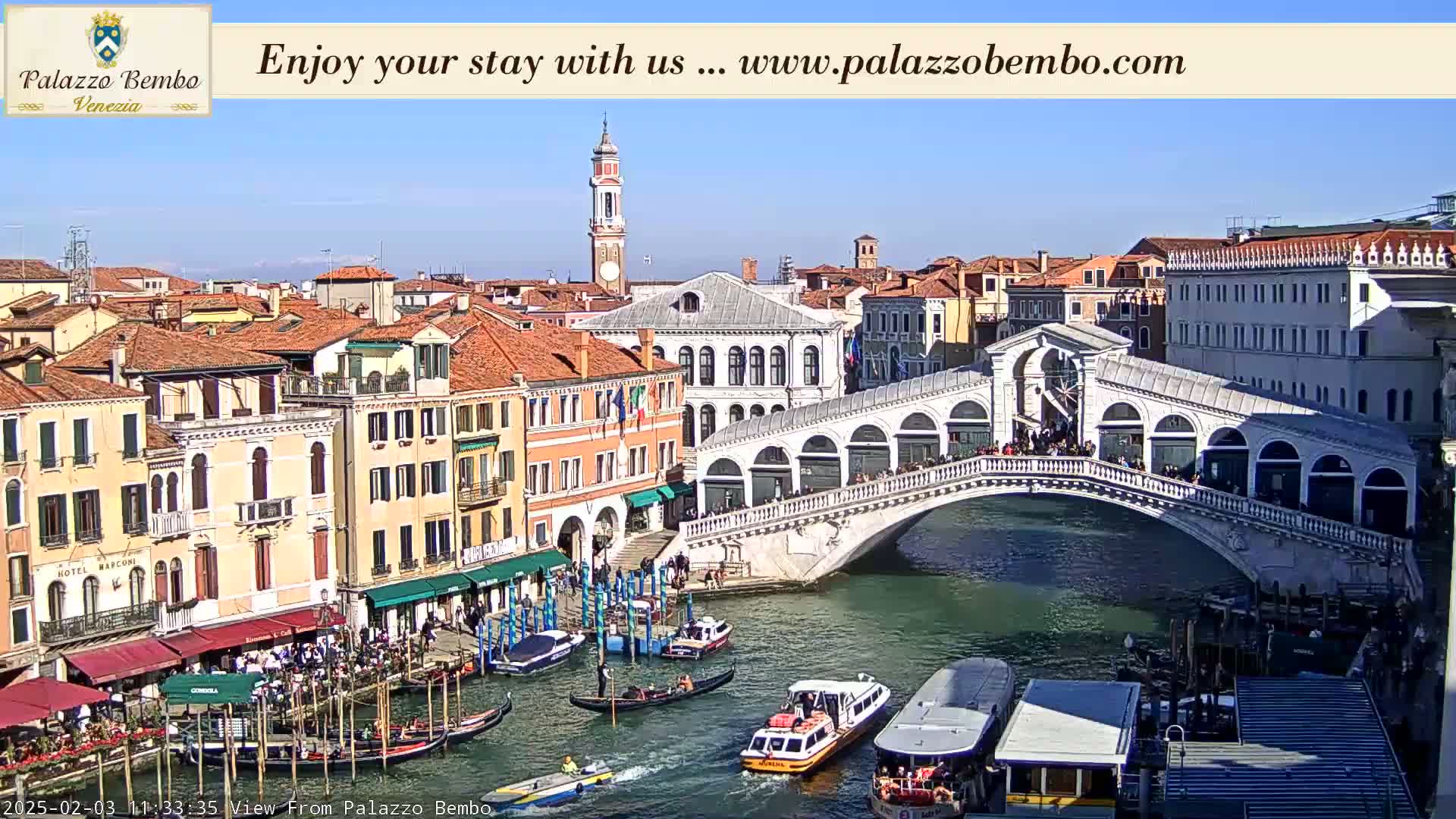 A sunny day in Venice shows a canal with gondolas and water taxis passing under a stone bridge lined with people, with colorful buildings on either side.