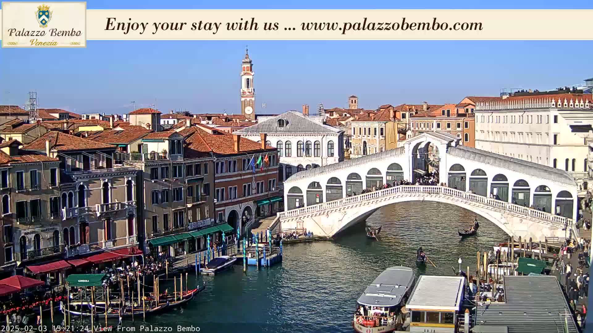 A sunny day in Venice shows a canal with gondolas and boats passing under a bridge lined with people, with buildings and rooftops lining the canal on both sides.
