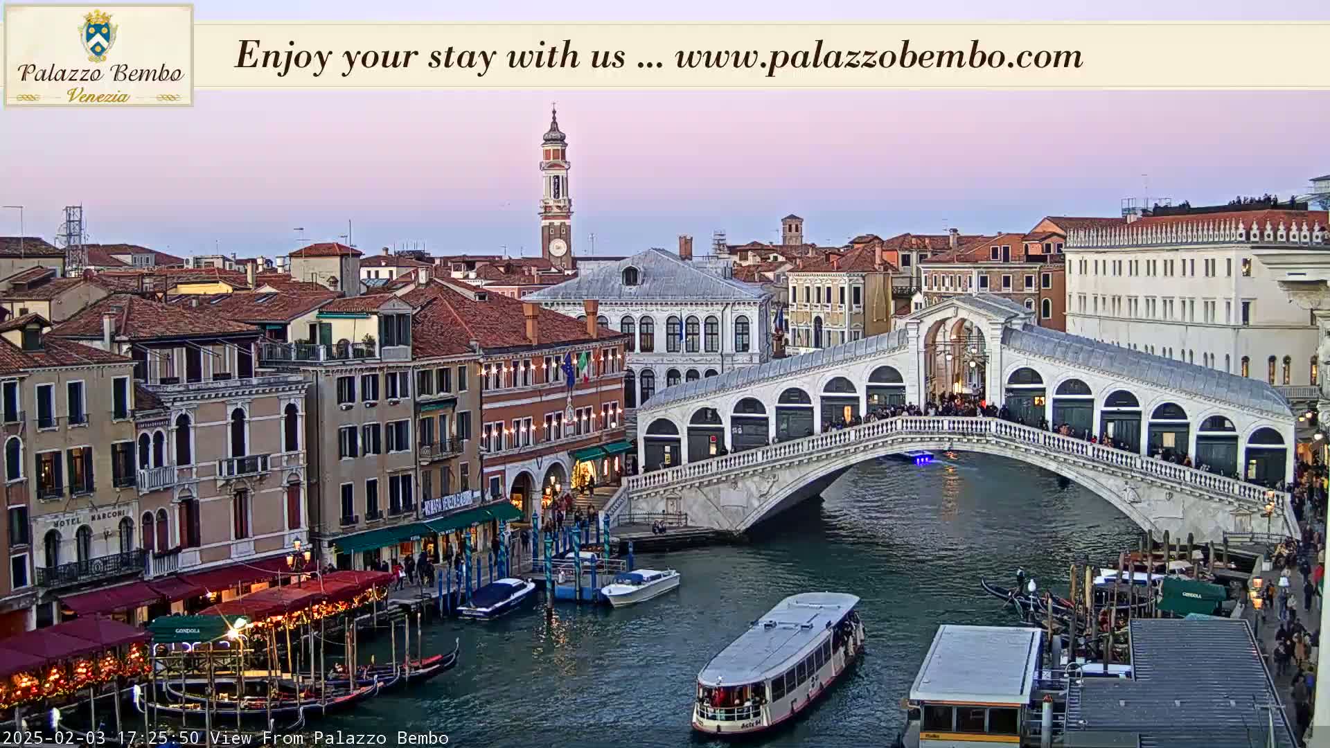 A twilight view of a Venetian canal, showing a bridge crowded with people, gondolas, boats, and buildings lining the waterway.