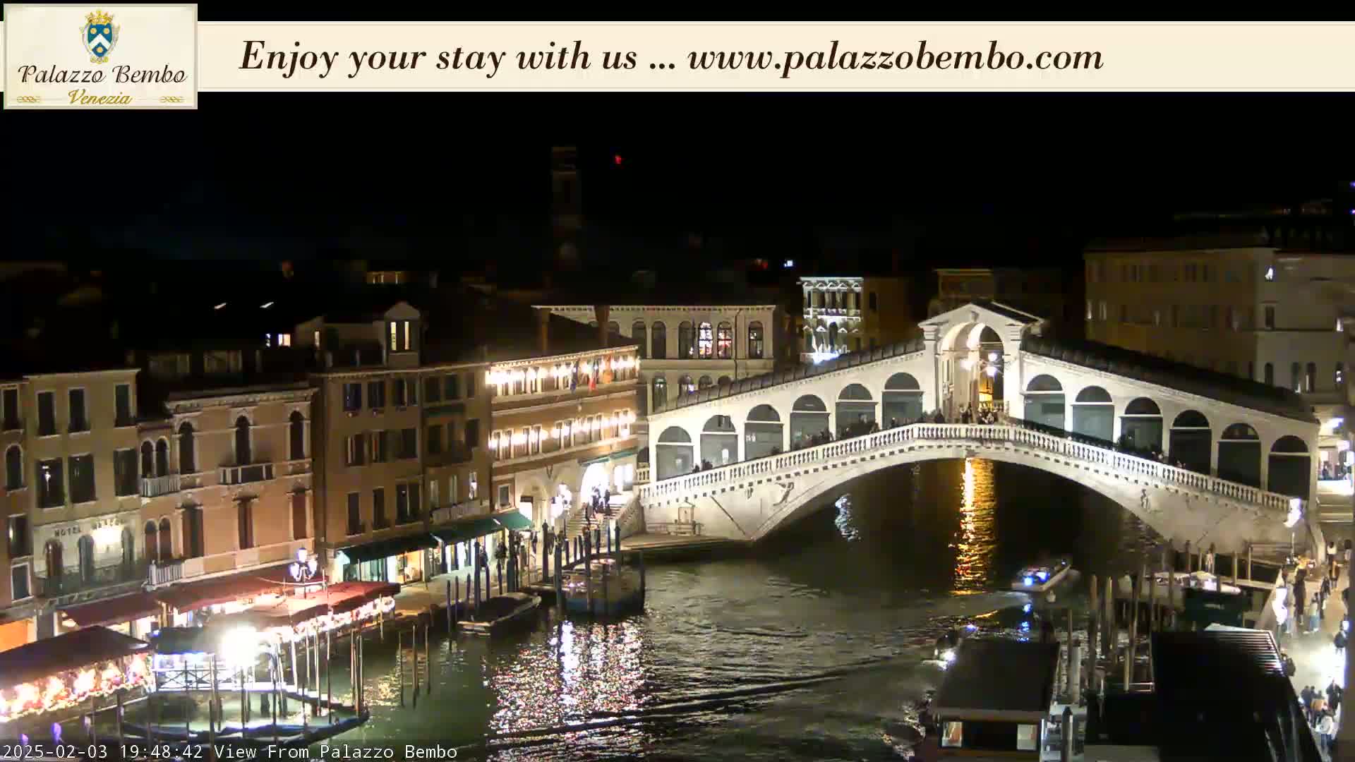 A nighttime view of a canal in Venice, showing the Rialto Bridge illuminated and lined with people, with buildings and boats along the water's edge.