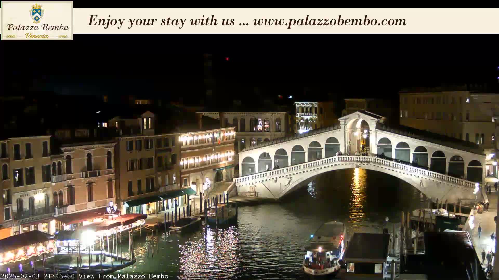 A nighttime view of a canal in Venice, showing a bridge lit up with lights reflecting on the water, and buildings along the waterway.