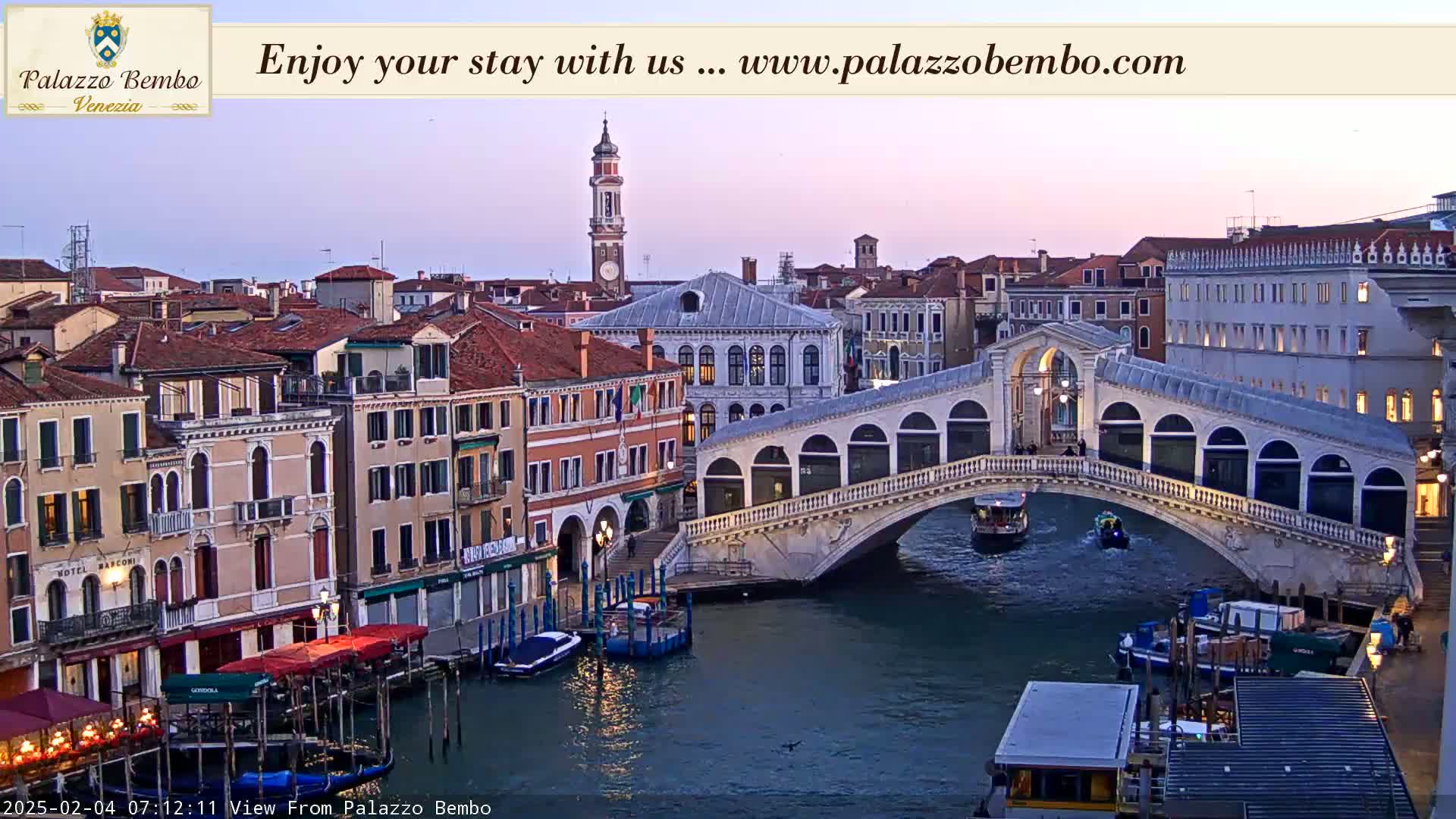 A twilight view of a canal in Venice, Italy, shows a bridge with gondolas and boats passing under it, with buildings lining the canal on either side.