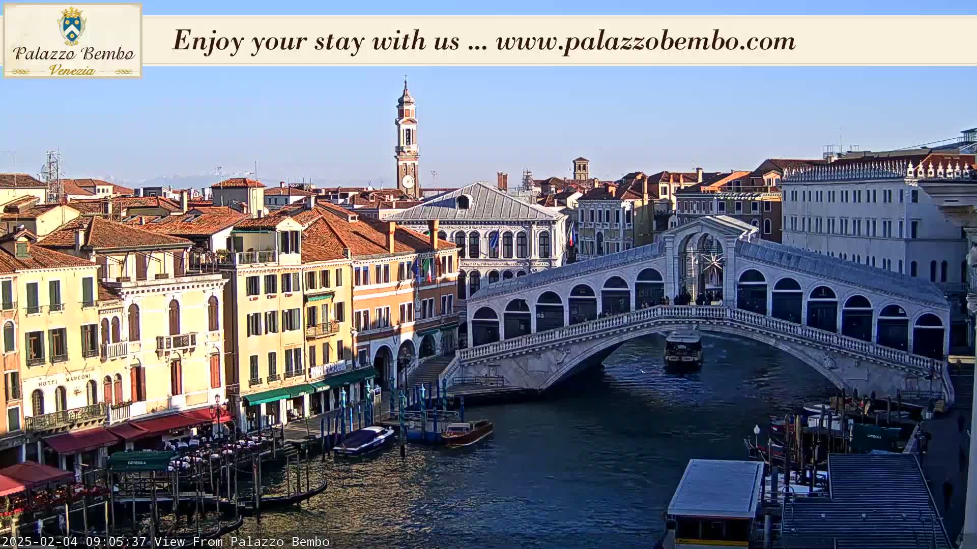 A sunny view of a canal in Venice, featuring the Rialto Bridge, gondolas, and numerous buildings along the waterway.