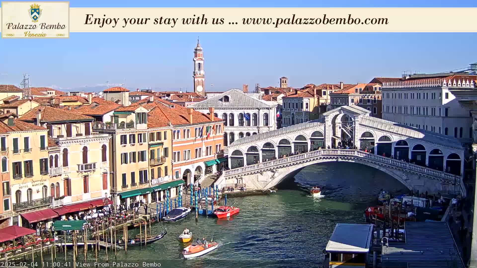 A sunny view of a Venetian canal, showing a bridge with people walking across it, boats in the water, and colorful buildings lining the canal.