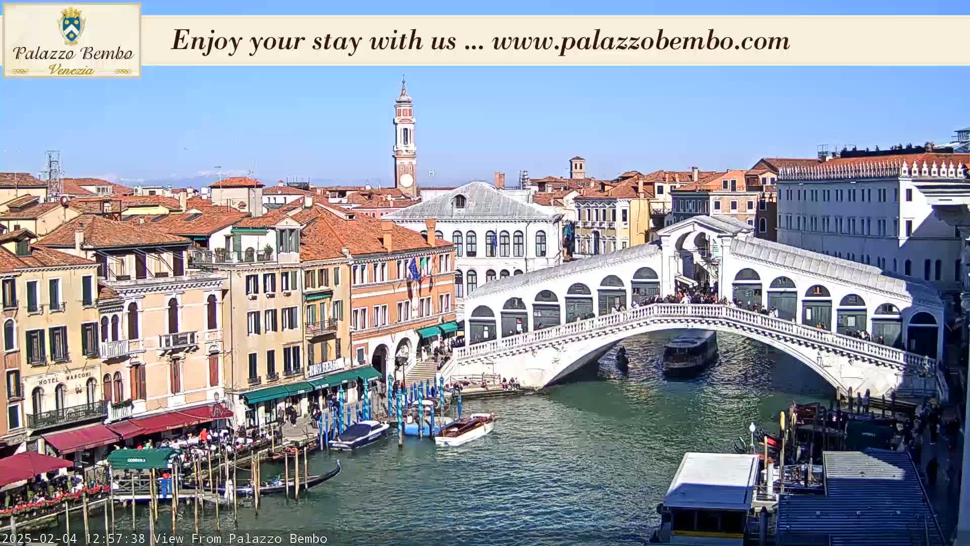 A sunny view of a canal in Venice, Italy, shows a bridge with people walking across it and boats moored along the canalsides.