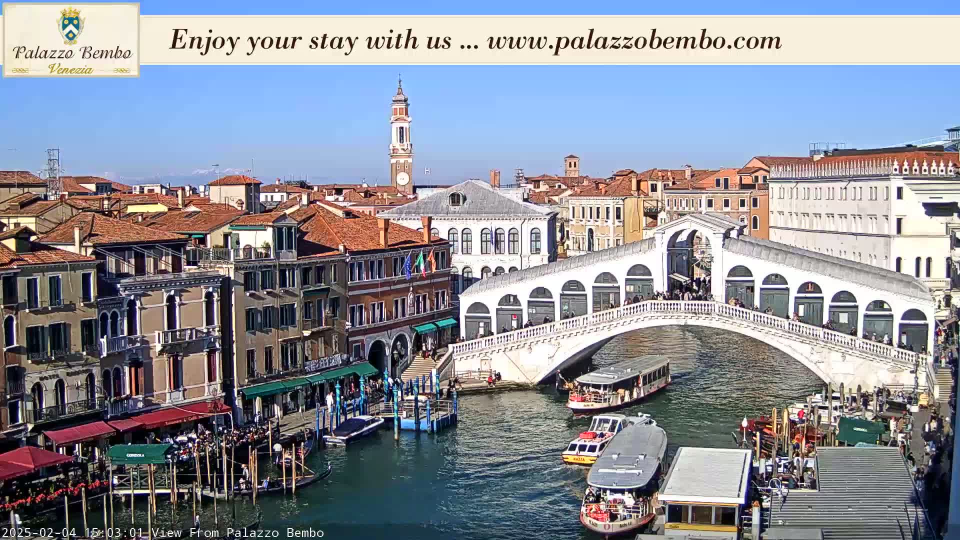 A sunny day in Venice shows a canal with boats, the Rialto Bridge, and numerous buildings lining the waterway.