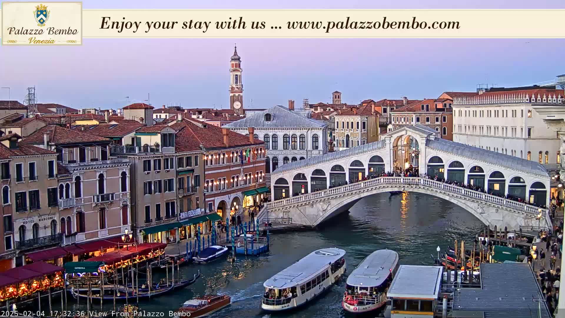 A twilight view of a canal in Venice, showing a bridge crowded with people and boats moored along the waterway, with buildings lining the canal.